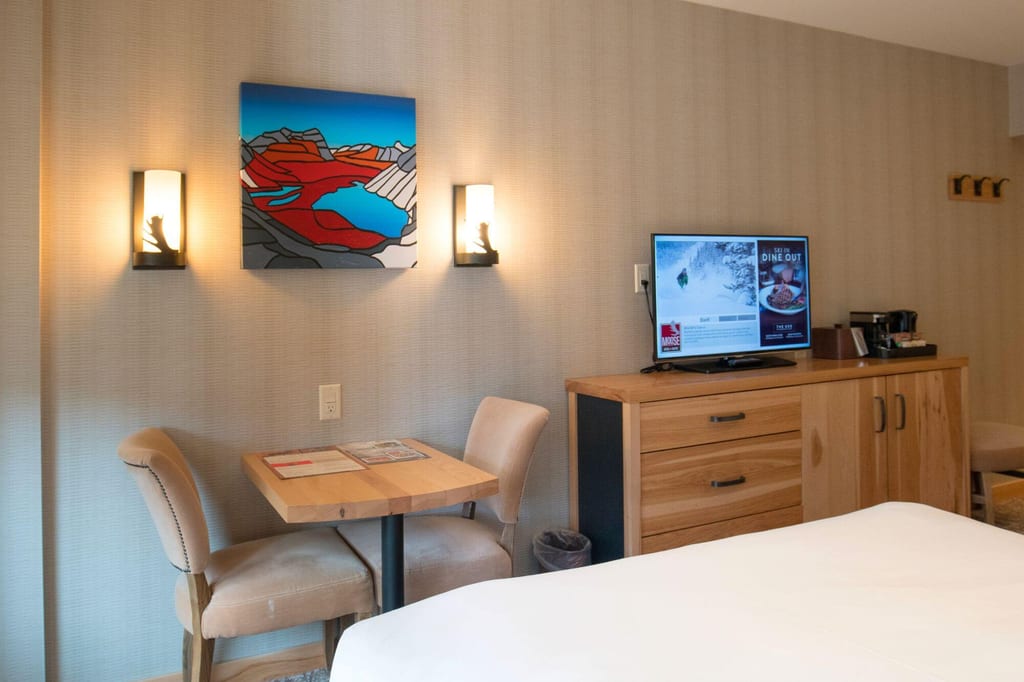 Hotelzimmer mit Tisch, zwei Stühlen, Wandbild und Sideboard mit Fernseher bei warmer Beleuchtung.