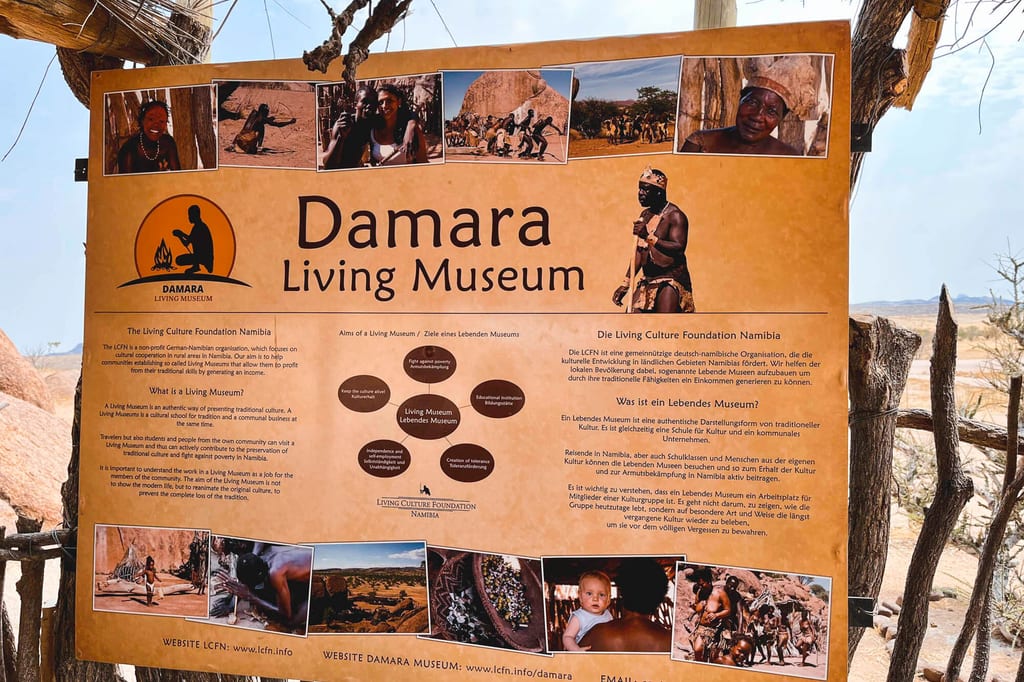 Info-Schild ‚Damara Living Museum‘ mit Bildern und trockener Landschaft im Hintergrund bei hellem Tageslicht.