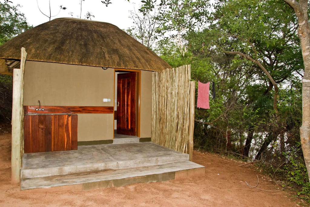 Strohdach-Rundhütte mit offener Holztür, Veranda und Waschbecken neben Buschland am Flussufer.