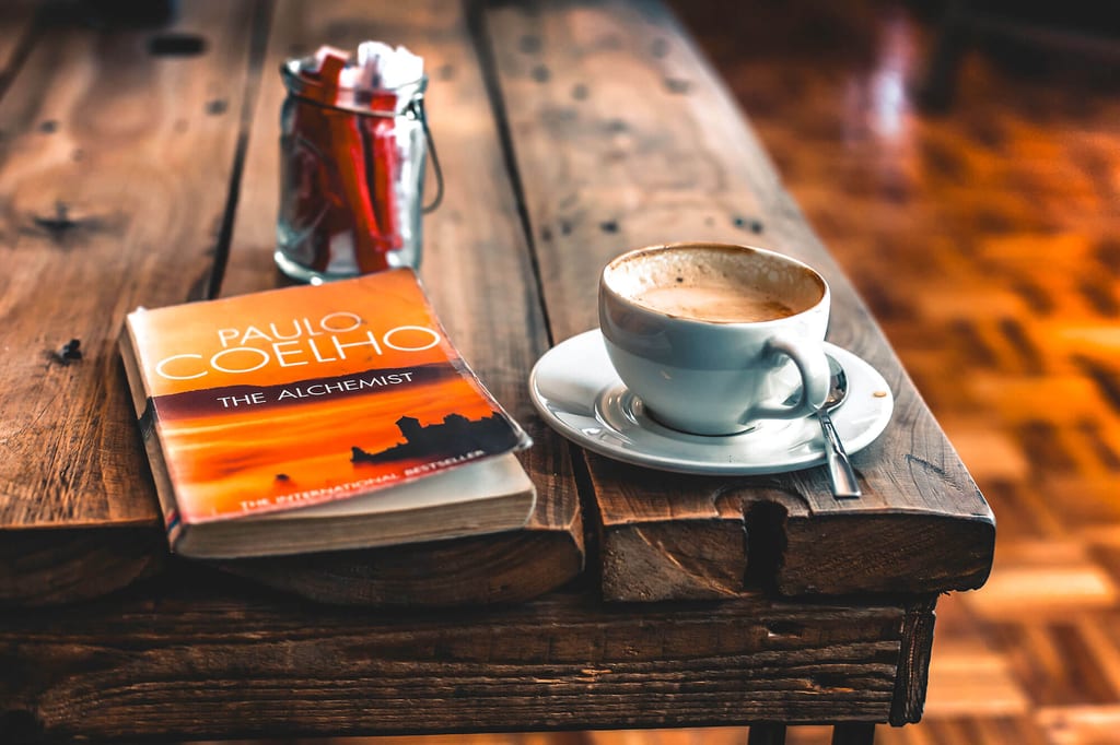 Taschenbuch ‚The Alchemist‘ und eine Tasse Kaffee auf rustikalem Holztisch in warmem Innenlicht.