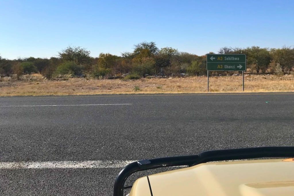 Straßenblick in Botswana mit Wegweiser nach Ghanzi, trockene Buschsavanne und Fahrzeugmotorhaube im Vordergrund.