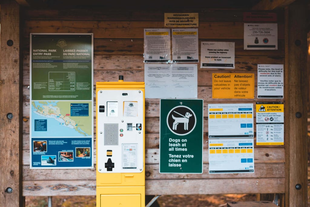 Frontale Aufnahme eines Holz-Anschlagbretts mit Informationsschildern und gelbem Parkautomaten.