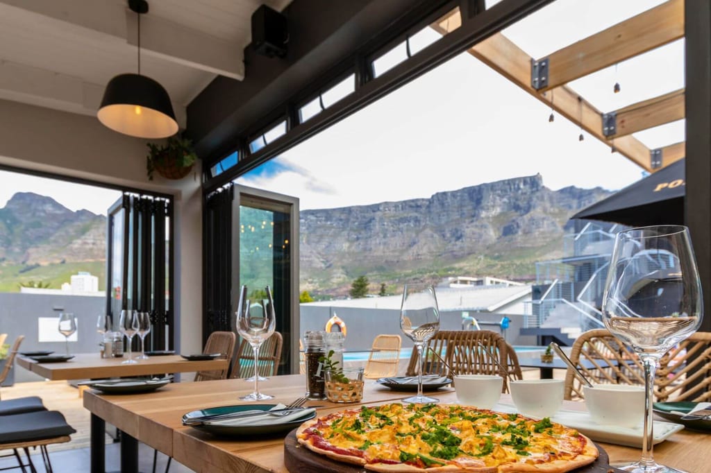 Gedeckter Holztisch mit Pizza und Gläsern, offene Türen und Bergblick am Tag