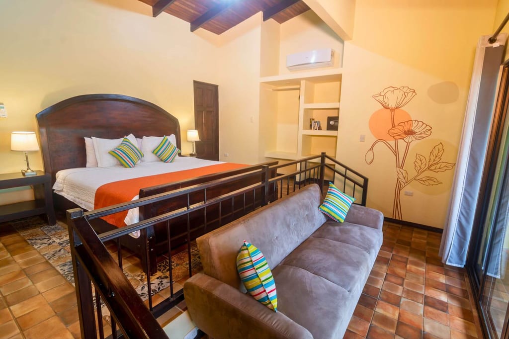 Helles Hotelzimmer mit Doppelbett, grauer Couch, bunten Kissen und Blumenwandmotiv