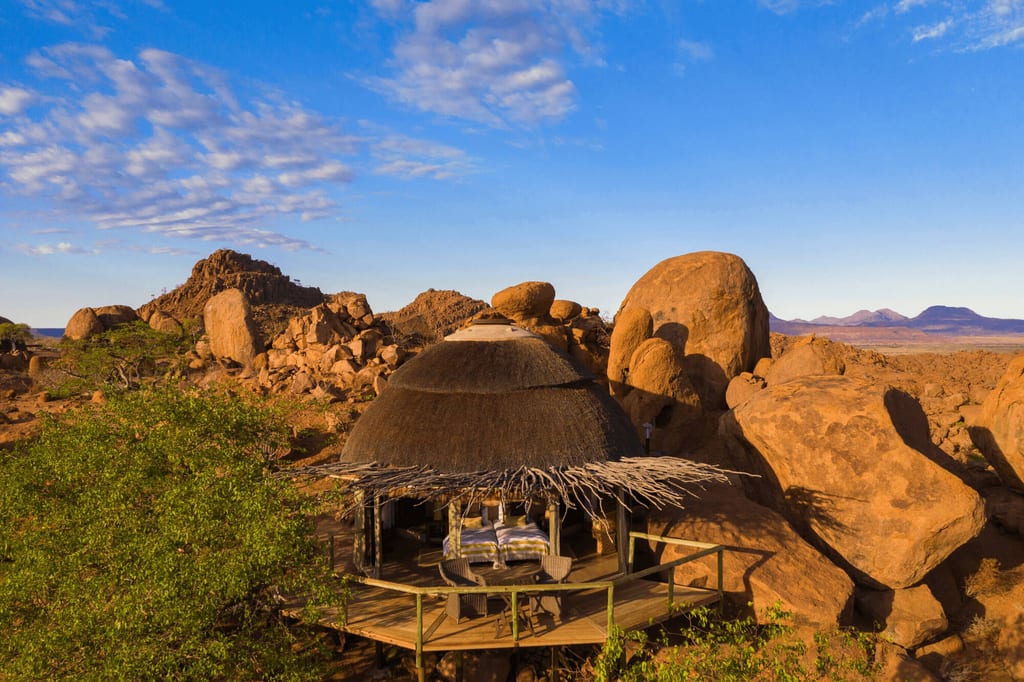 Runde Strohlodge auf Terrasse zwischen roten Felsen im warmen Abendlicht, weiter Blick über trockenes Plateau.