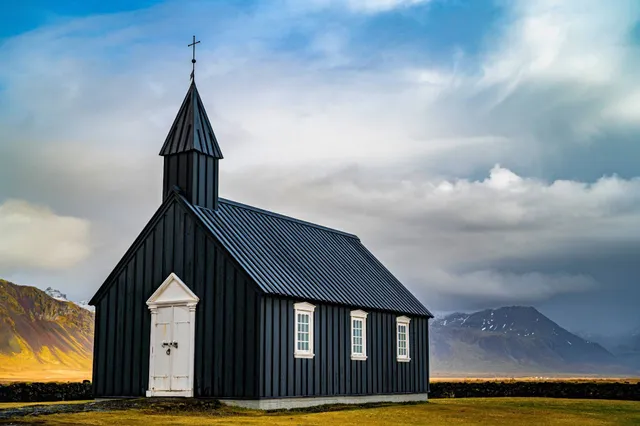Schwarze Holzkirche vor Bergen und dramatischem Wolkenhimmel in karger Landschaft.