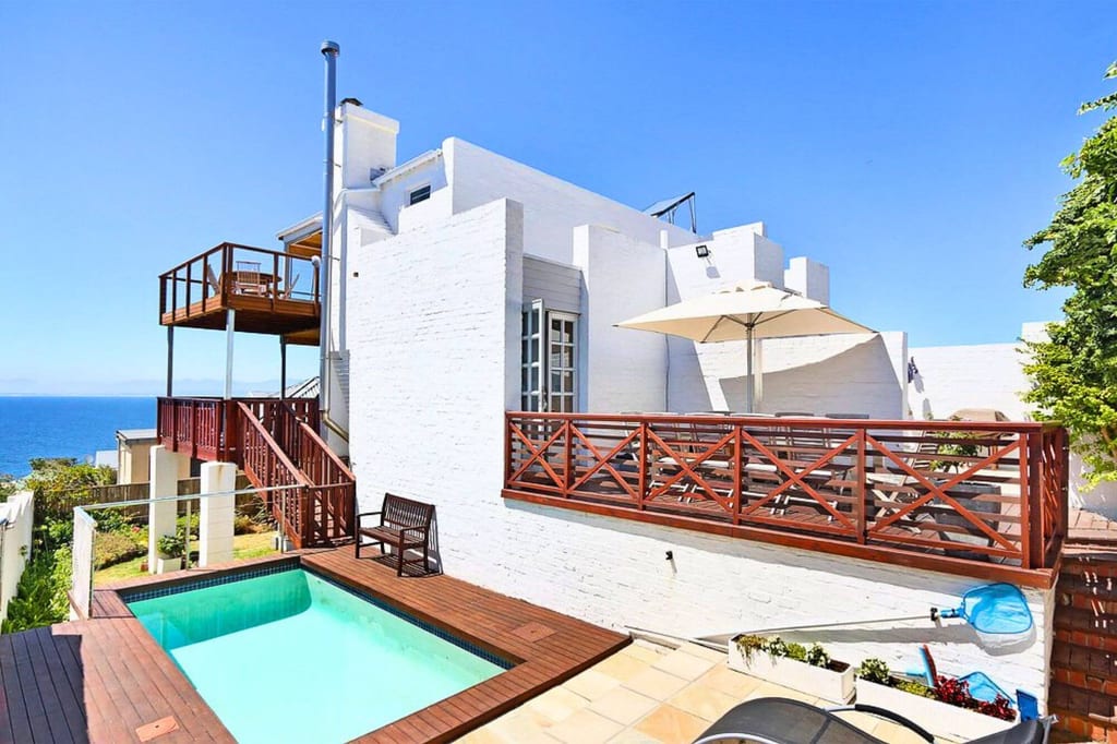 Weißes Küstenhaus mit Holzterrassen, kleinem Pool und Meerblick bei strahlendem Sonnenschein.