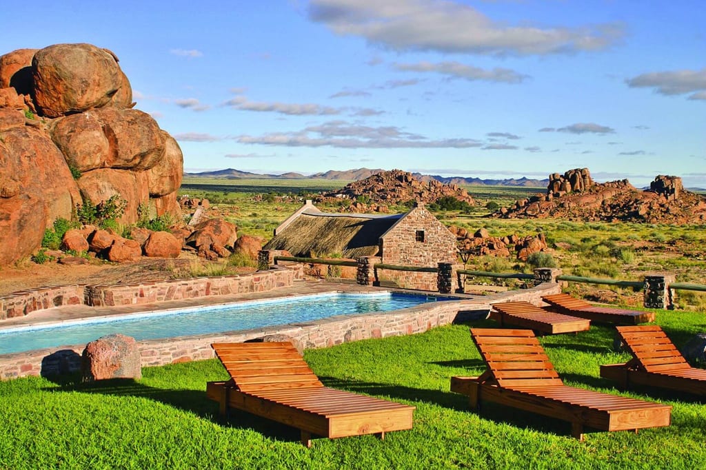 Pool mit Holzliegen auf Rasen vor kleinem Steinhaus und roten Felsen in weitem Grasland
