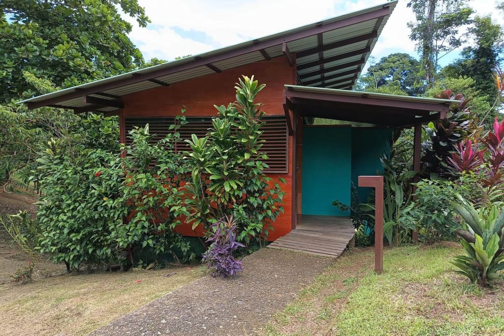 Kleines rotes Holzhaus mit überdachter Veranda, umgeben von dichtem tropischem Grün und einem Kiesweg.