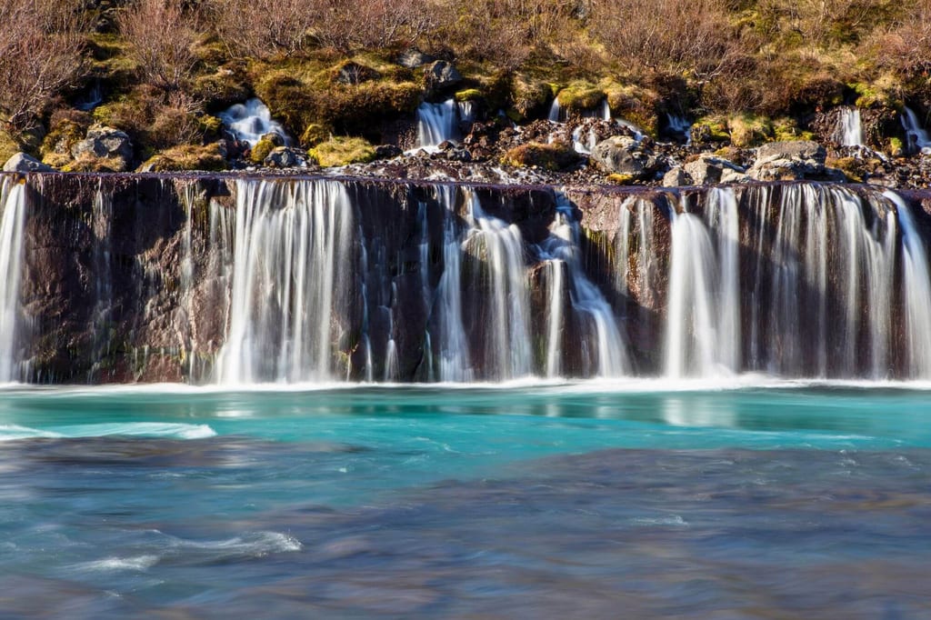 Mehrere schmale Wasserfälle stürzen über eine dunkle Felsstufe in ein türkisfarbenes Flussbecken.