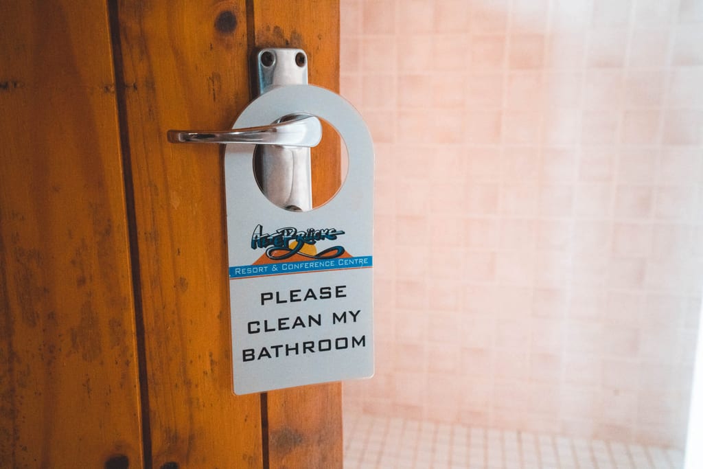 Türschild mit der Aufschrift ‚PLEASE CLEAN MY BATHROOM‘ hängt an einem Türgriff vor gefliestem Bad