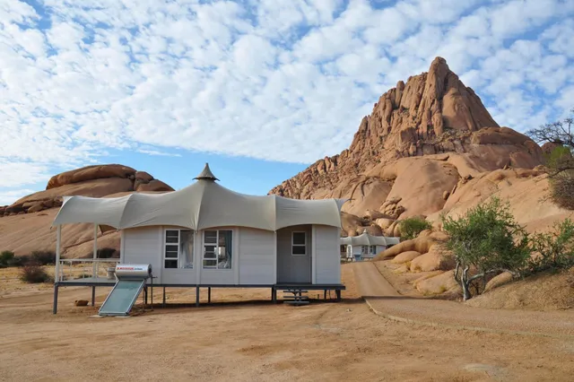 Safari-Bungalows vor einem markanten Granitfelsen in trockener, sonniger Landschaft mit Fleckenwolken