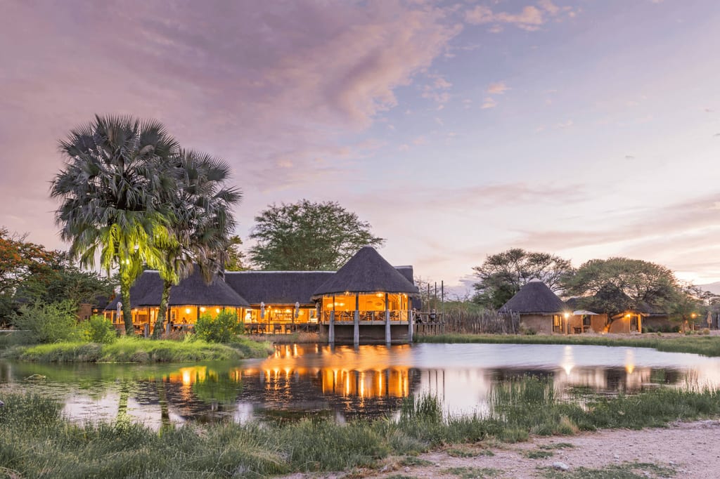Beleuchtete Lodge mit Strohdächern spiegelt sich bei Abenddämmerung in ruhigem Wasser.