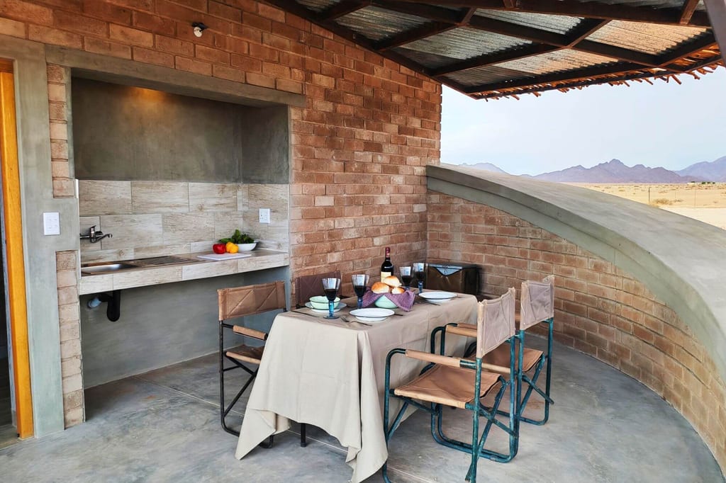 Überdachte Terrasse mit gedecktem Tisch, Küchennische und Blick auf trockene Landschaft und Berge im Abendlicht.