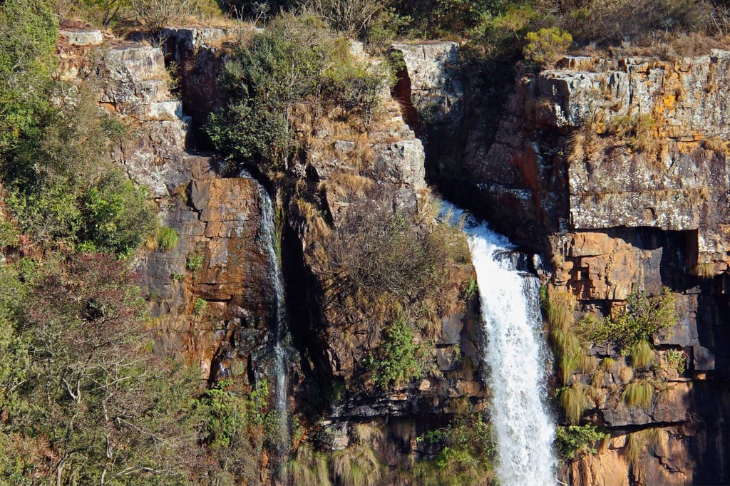 Schmaler Wasserfall stürzt an einer sonnenbeschienenen Felswand neben trockenem Gras und Sträuchern hinab.