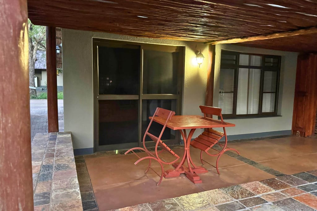 Überdachte Veranda mit rotem Tisch und zwei Stühlen, abendliche Beleuchtung durch Wandlampe.