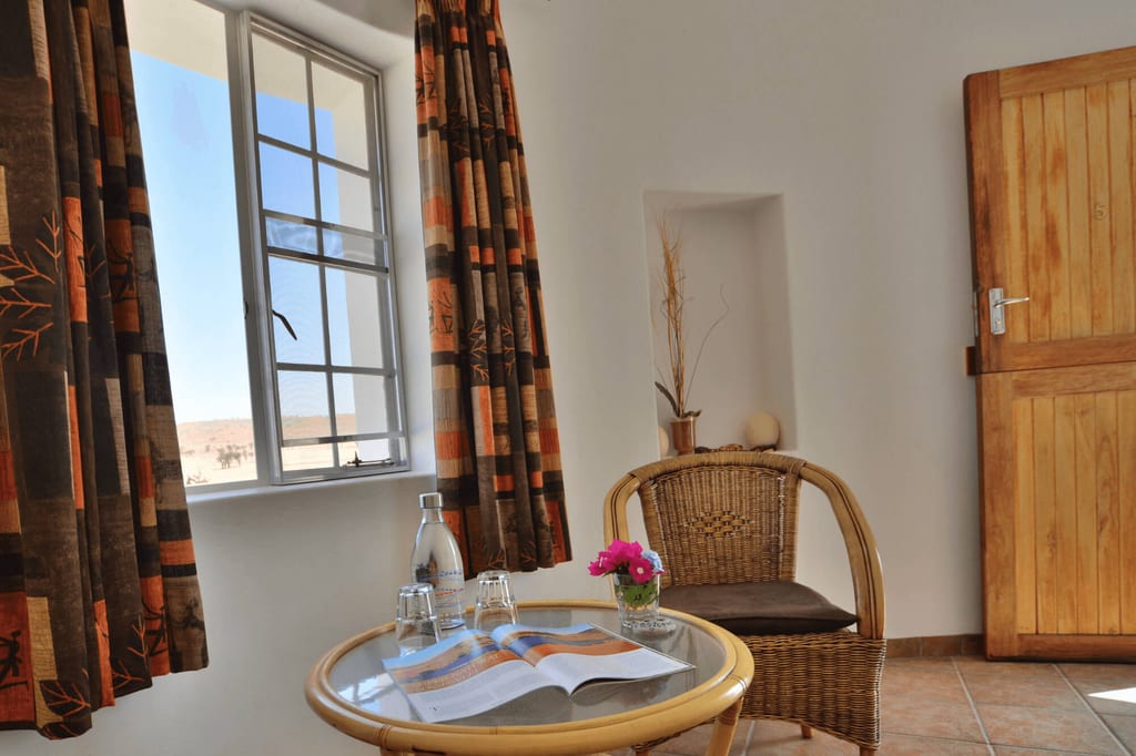 Helles Zimmer mit Rattanstuhl, rundem Glastisch und offenem Fenster mit Blick auf sandige Landschaft.