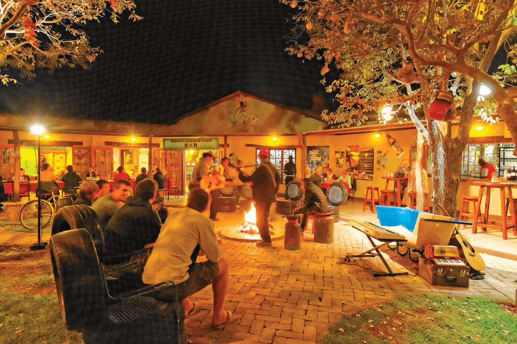 Abendliche Bar mit Gästen um ein offenes Feuer, Musiker spielen Gitarre, warme Lichtstimmung.