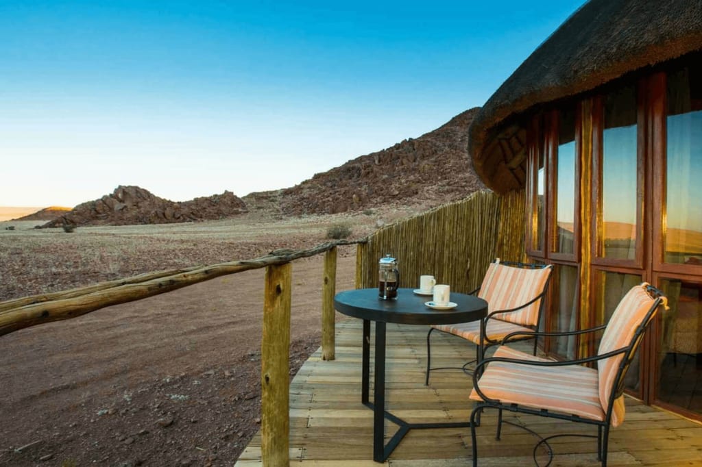 Holzveranda mit zwei Stühlen und Kaffeetisch vor felsiger Halbwüstenlandschaft im warmen Seitenlicht.