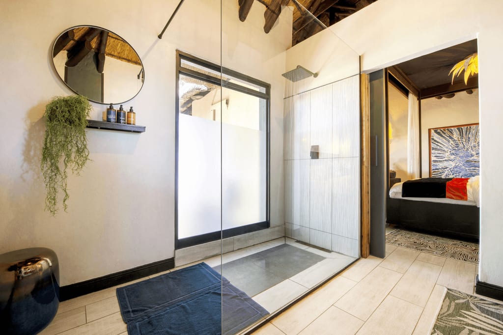 Helles Badezimmer mit Glasdusche, rundem Spiegel und Blick in ein angrenzendes Schlafzimmer.