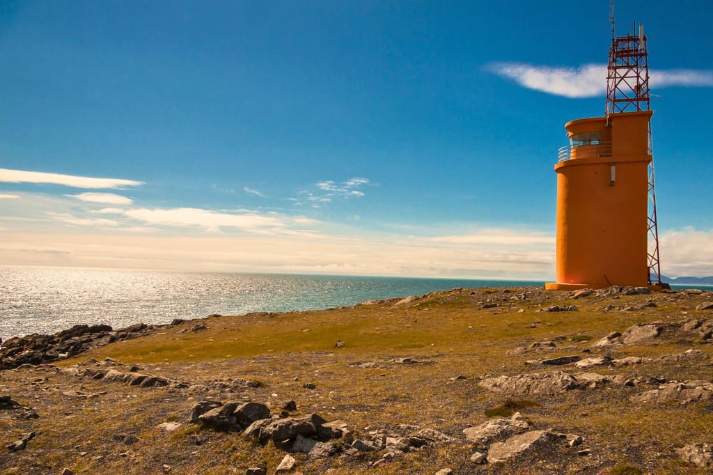 Orangefarbene Küstenstation auf felsiger Landzunge mit Meer und blauem Himmel bei Sonnenschein.