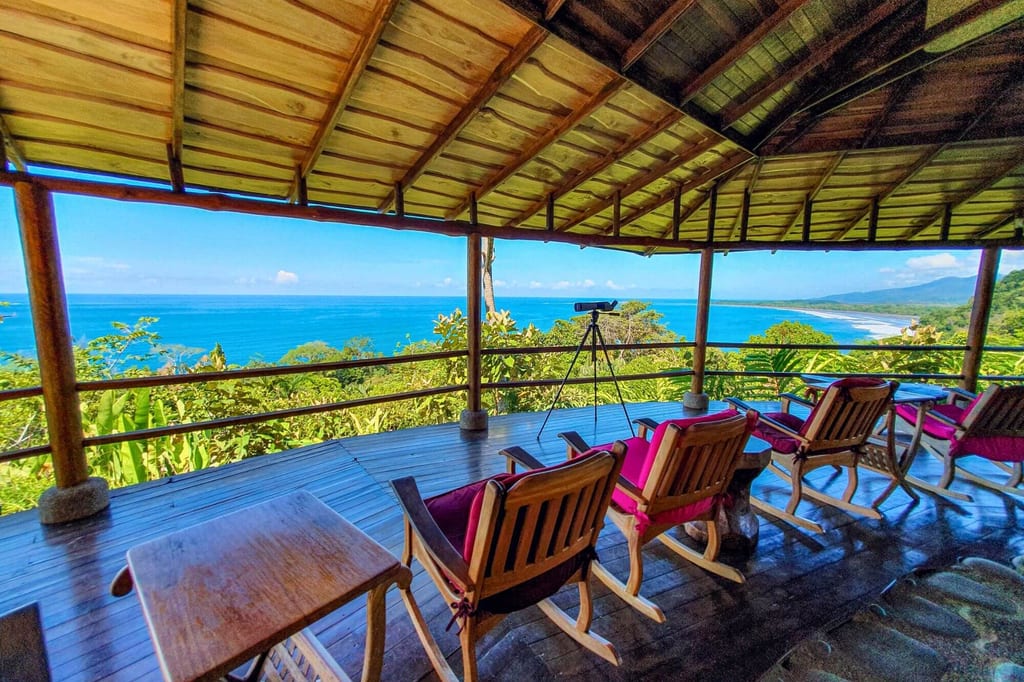 Überdachte Holzterrasse mit Stühlen und Fernglas, Aussicht auf blaues Meer und grüne Küste
