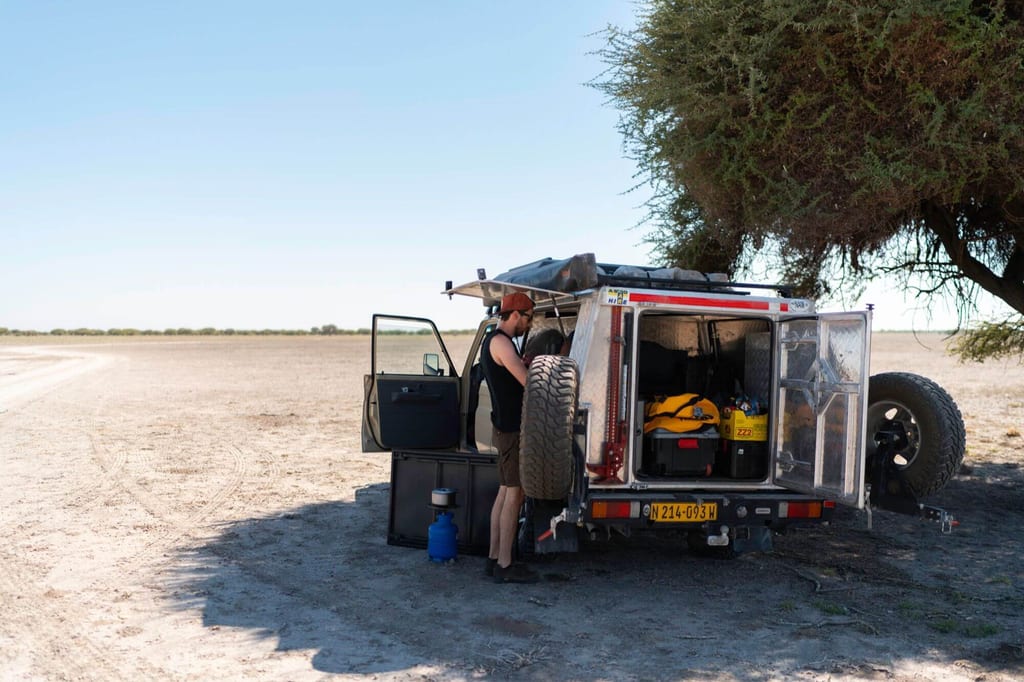 Person steht am offenen Heckanhänger eines Offroad-Campers im Schatten eines Baums auf trockener Ebene.
