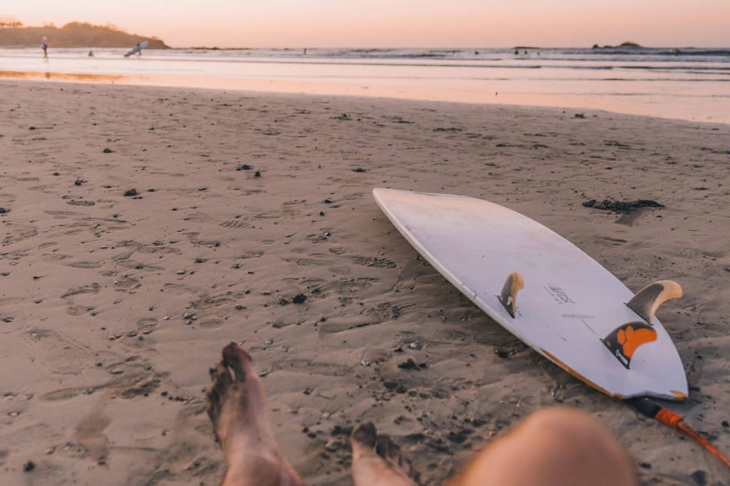 Surfboard liegt am Strand, verschwommene Beine im Vordergrund, ruhiges Meer im Abendlicht