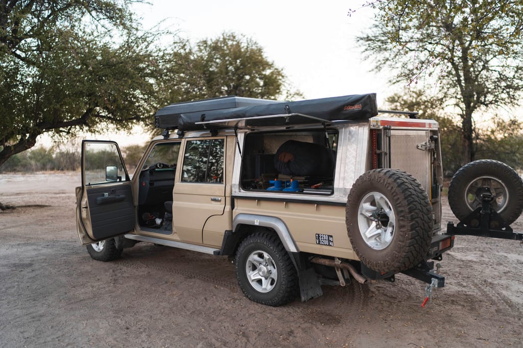 Beiges Offroad-Campingfahrzeug mit geöffneten Türen und Dachzelt unter Bäumen im Abendlicht.