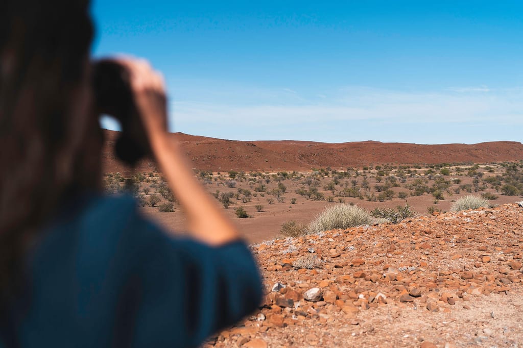 Blick über die Schulter einer Person mit Fernglas auf rote, steinige Wüstenlandschaft unter klarem Himmel.