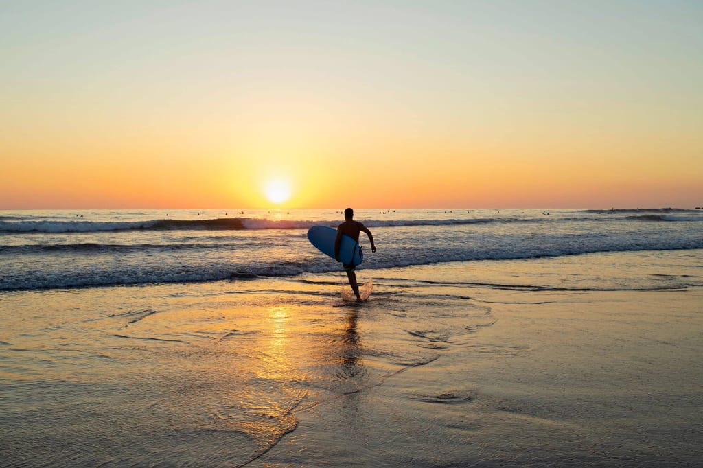 Surfer als Silhouette mit Surfboard läuft bei Sonnenuntergang ins Meer, Reflexe auf nassem Sand.