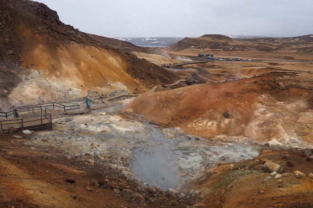 Geothermisches, dampfendes Gebiet mit bunten Hügeln und Holzstegen bei bewölktem Himmel.
