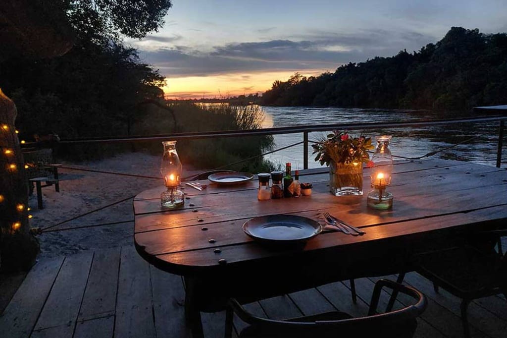 Abendlicher Flussblick mit gedecktem Holztisch, Öllampen und Blumen im warmen Licht des Sonnenuntergangs.