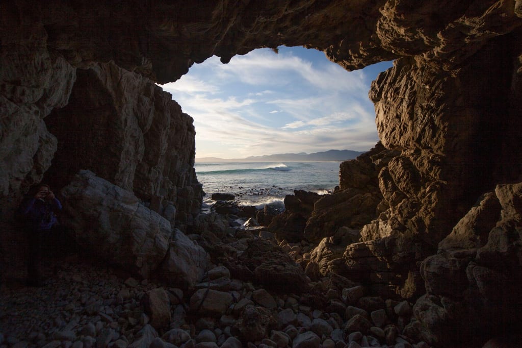 Blick aus einer Meereshöhle auf Wellen und ferne Berge im warmen Abendlicht.