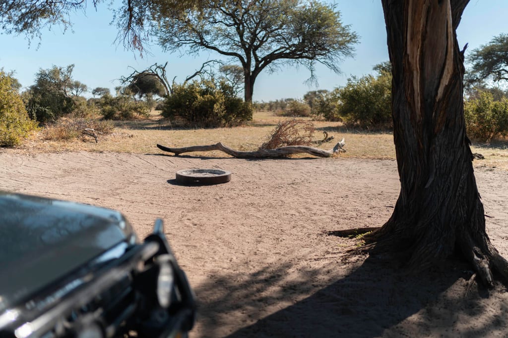 Blick neben Safari-Fahrzeug auf sandige Savanne mit Baum und metallischer Feuerstelle