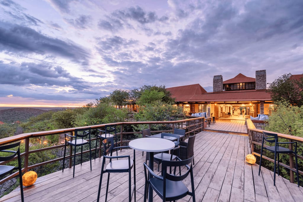 Holzterrasse mit Tischen und Stühlen vor einer warm beleuchteten Lodge bei Dämmerung