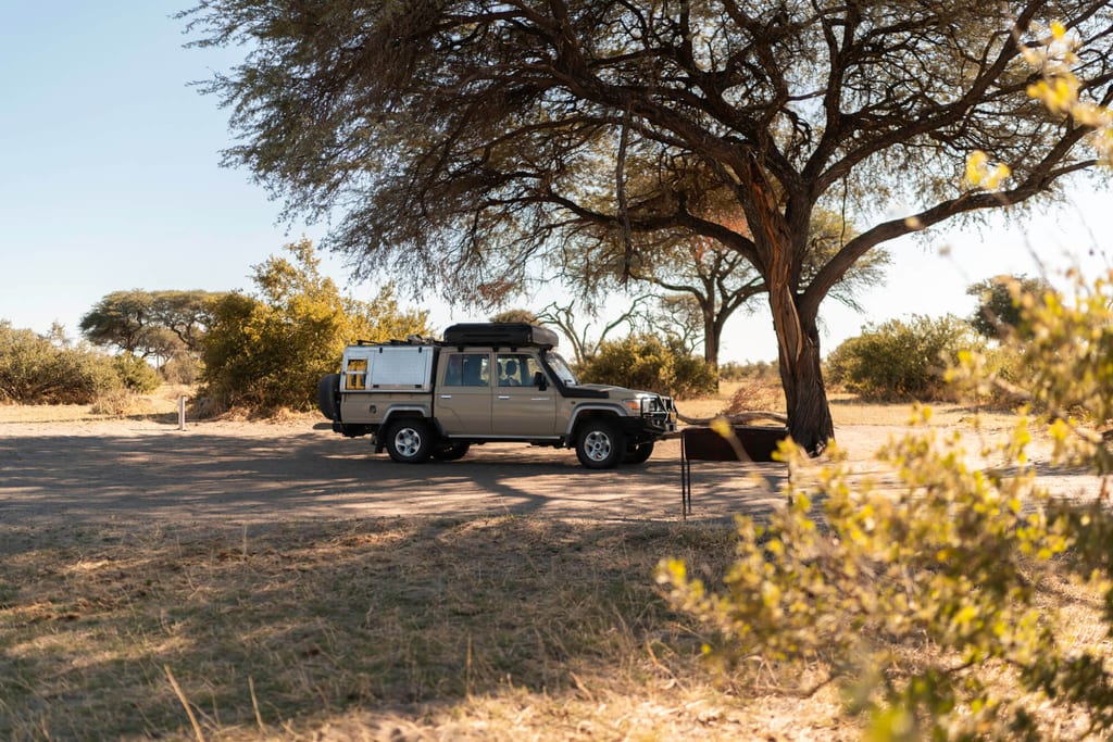 Geländewagen steht unter großem Baum in trockener Savannenlandschaft im warmen Nachmittagslicht.