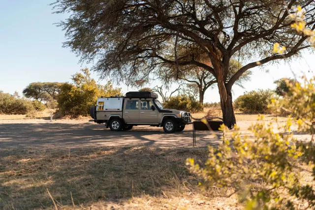 Geländewagen steht unter großem Baum in trockener Savannenlandschaft im warmen Nachmittagslicht.