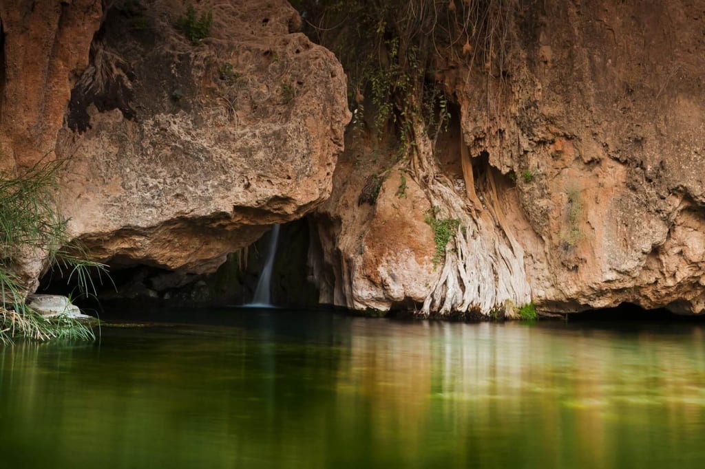 Kleine Höhle mit Wasserfall, stiller grüner Tümpel und verwitterte Felswand im weichen Licht.