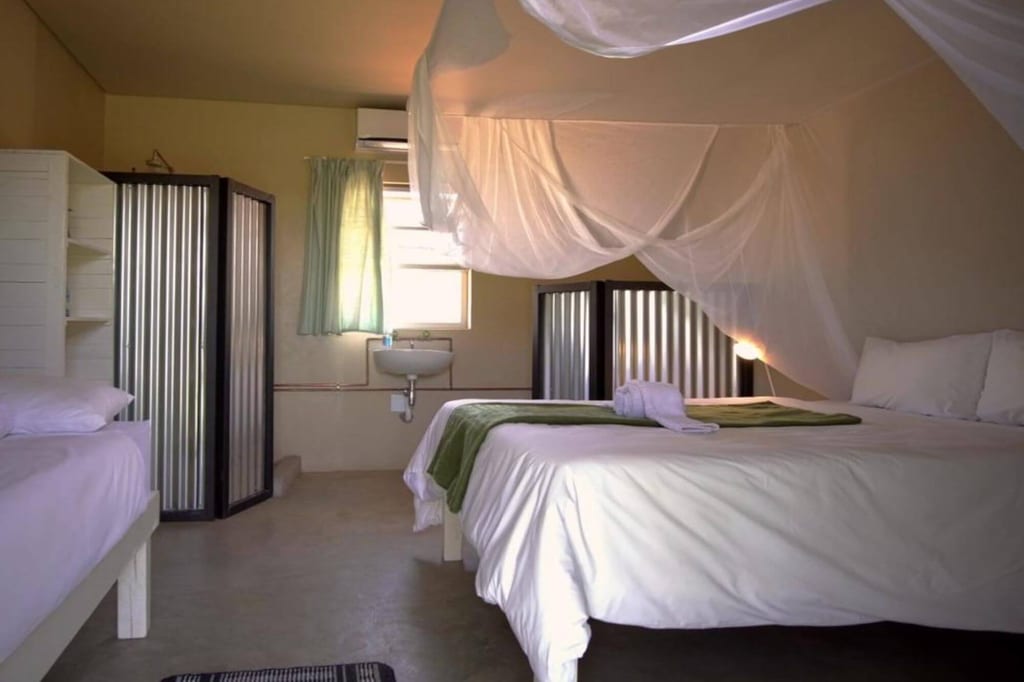 Schlichtes Gästezimmer mit Doppelbett, Moskitonetz und kleinem Waschbecken im Tageslicht.