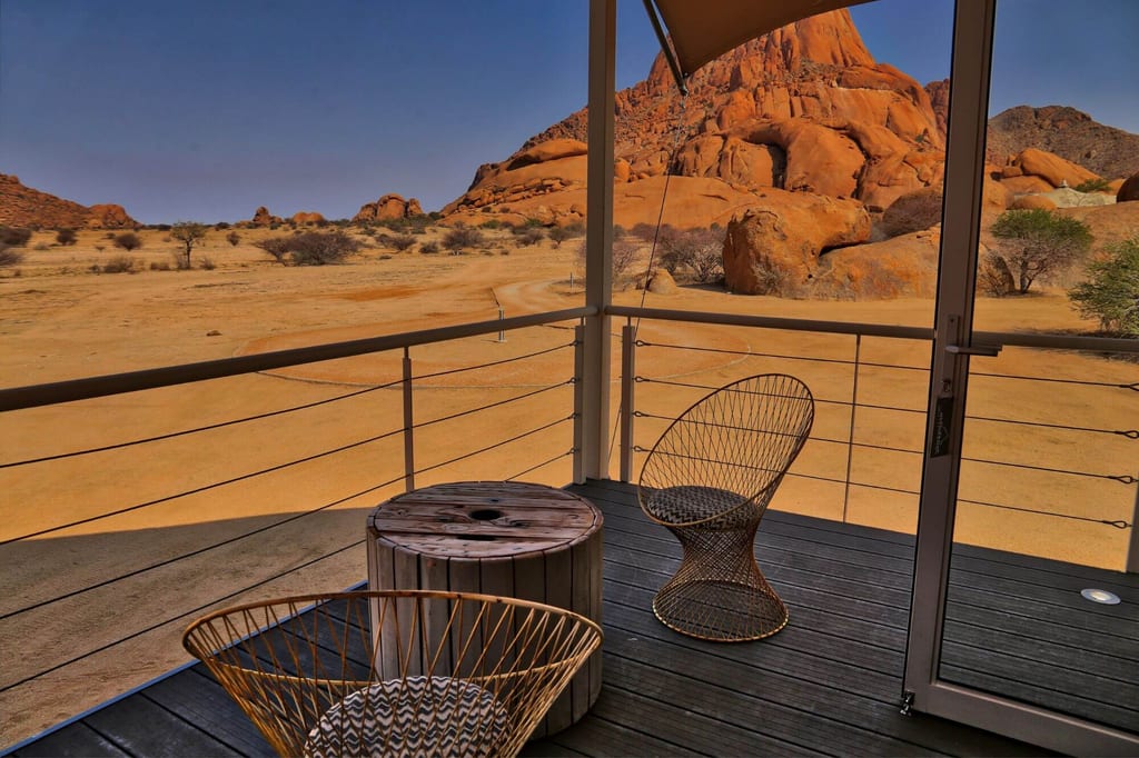 Terrasse mit Drahtstühlen und Blick auf orange Felsformationen in weiter Wüstenlandschaft bei warmem Licht.