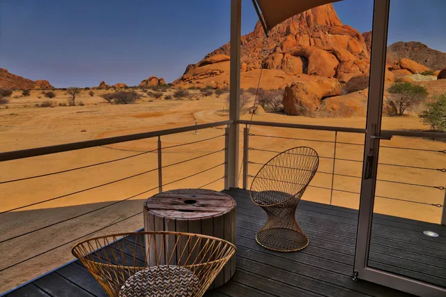 Terrasse mit Drahtstühlen und Blick auf orange Felsformationen in weiter Wüstenlandschaft bei warmem Licht.