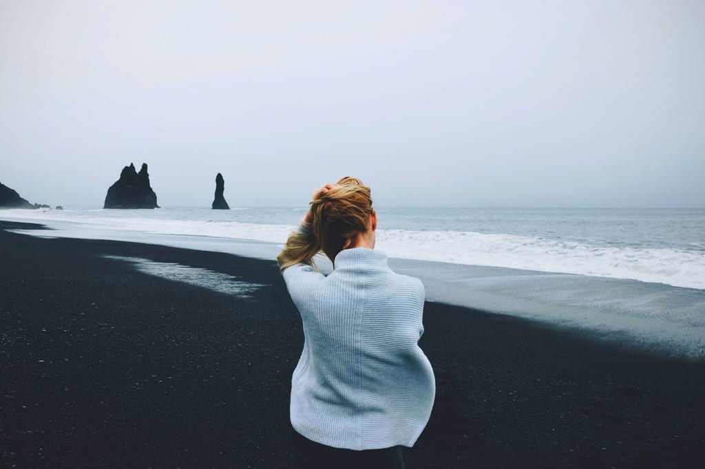 Person von hinten am schwarzen Sandstrand in Südisland, Meer und Felsnadeln bei bedecktem Himmel.