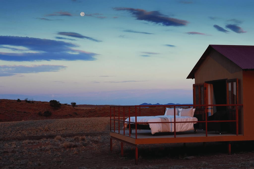 Bett auf Veranda einer Hütte in weiter, trockener Landschaft bei Dämmerung und mit sichtbarem Mond.