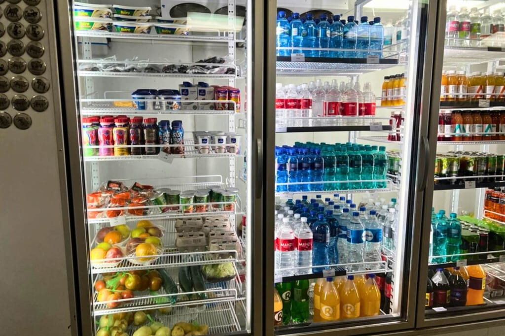 Frontalaufnahme eines beleuchteten Kühlregals mit Wasserflaschen, Säften, Obst und Eiern.