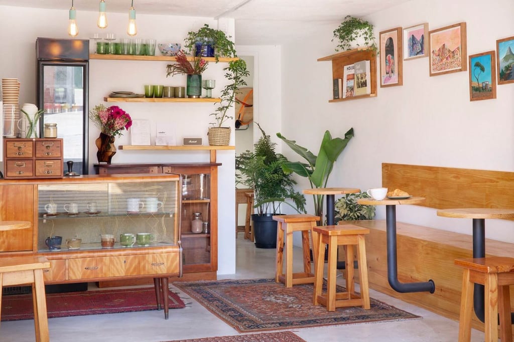 Helles gemütliches Café mit Holzmöbeln, Vitrine mit Tassen und Pflanzen an weißen Wänden.