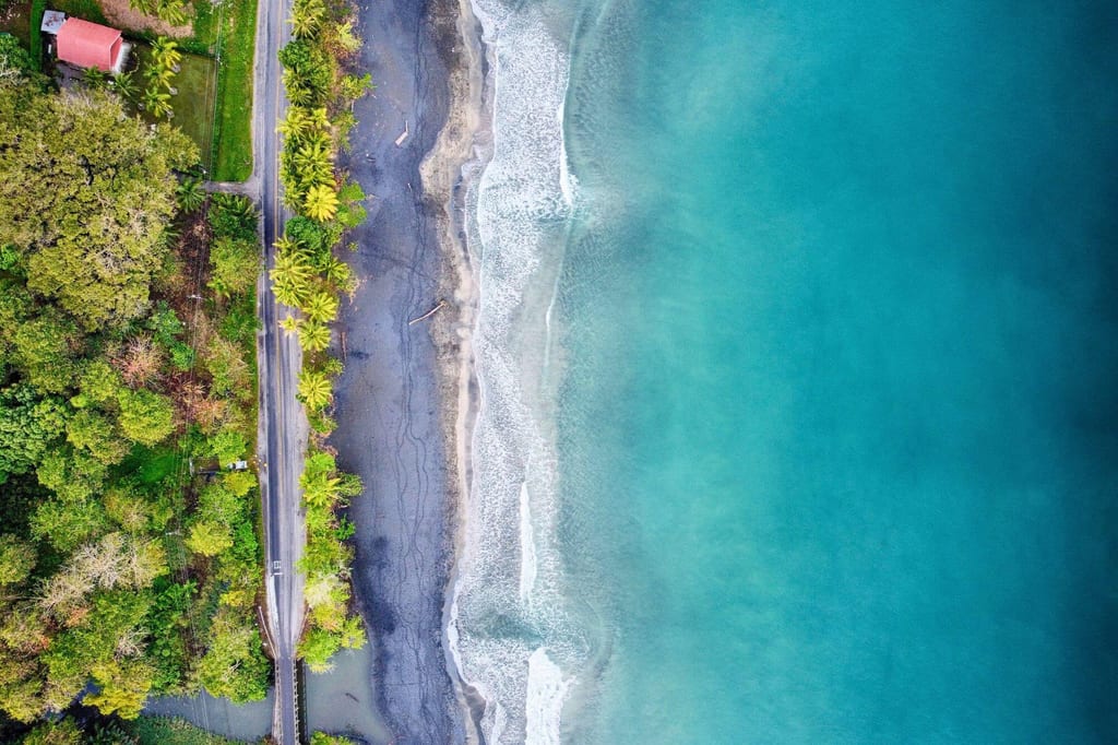 Luftaufnahme einer Küste mit türkisfarbenem Meer, dunklem Strand, Palmen und einer Straße