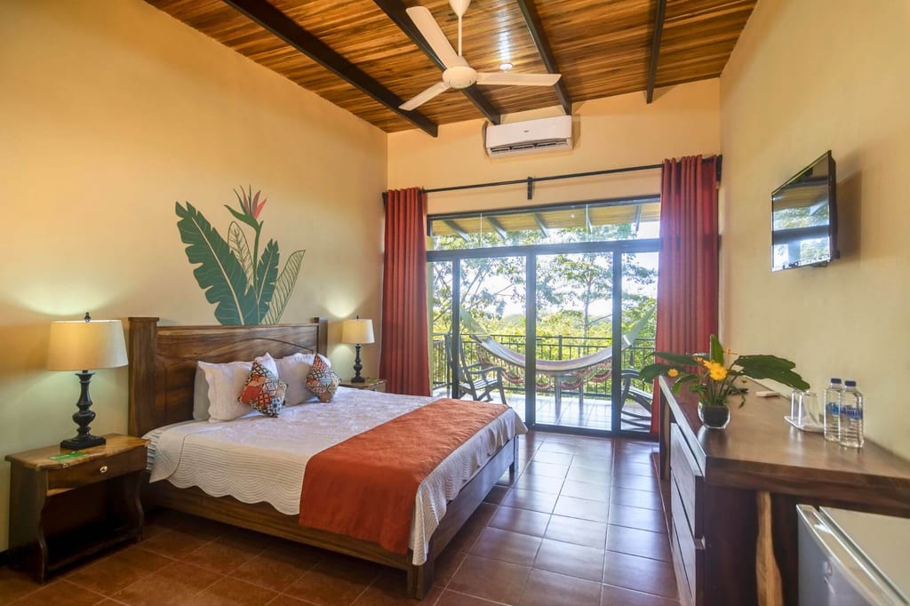 Helles Schlafzimmer mit Doppelbett und Balkon, Blick auf Hängematte und Grün draußen.