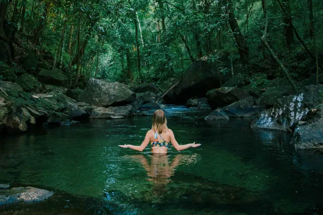 Person von hinten steht hüfttief in einem smaragdgrünen Waldfluss, umgeben von Felsen und dichter Vegetation.