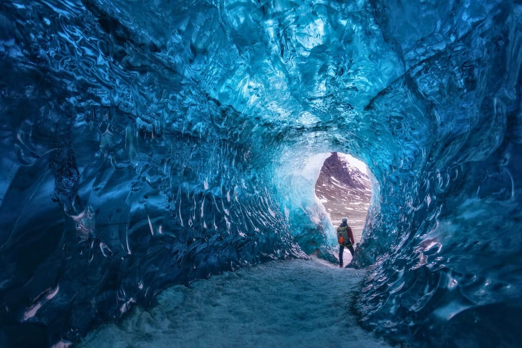 Blick aus einer blauen Eishöhle auf eine Person am Höhlenausgang bei Tageslicht.